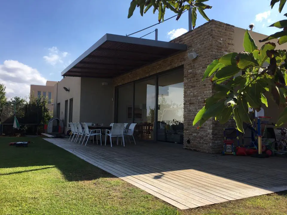 פרגולת אלומיניום תלויה צמודה לבית עם חצר ירוקה - עולם הפרגולות